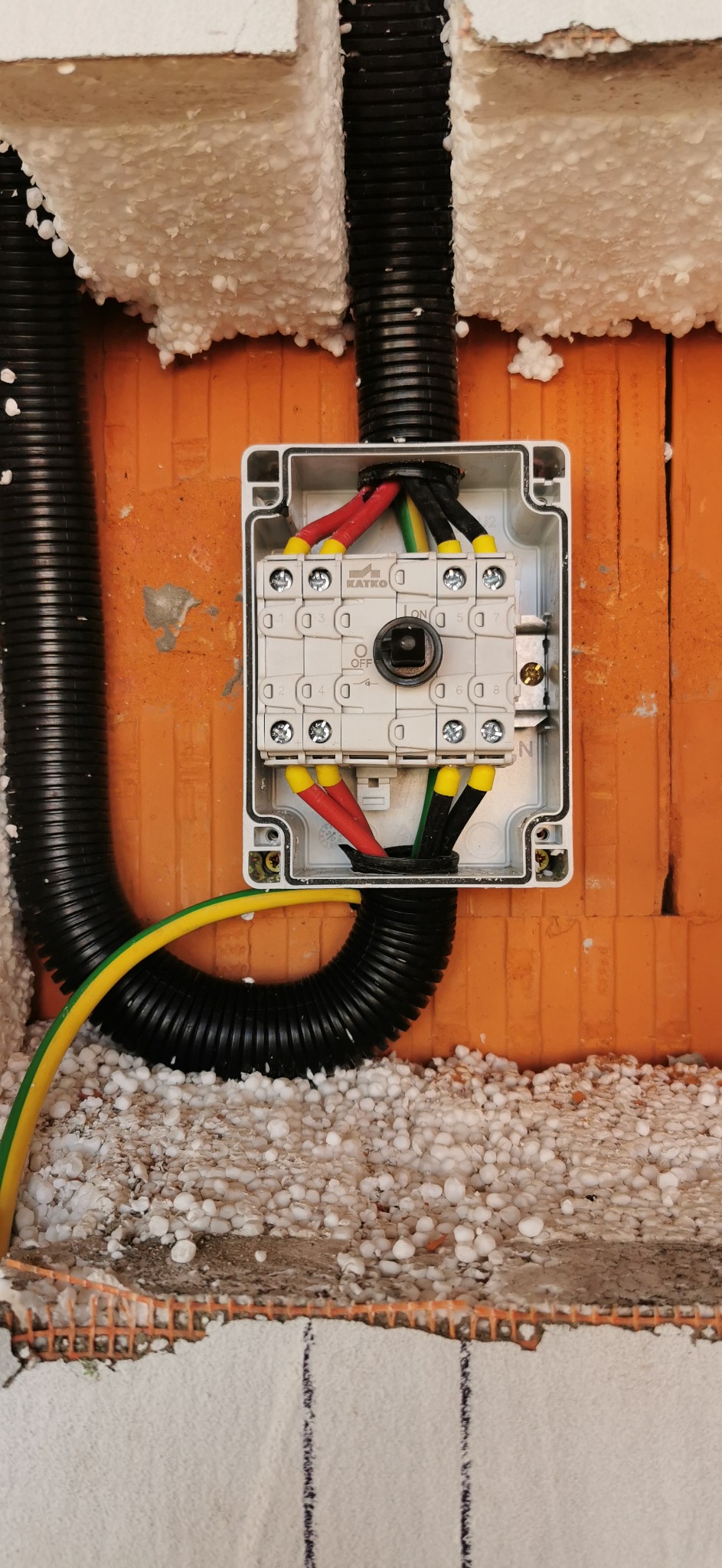 Rozdzielnica elektryczna Katko umieszczona w ceglanej ścianie z widocznymi przewodami w kolorach czerwonym, czarnym i zielono-żółtym, otoczona czarnymi rurkami karbowanymi i ociepleniem ze styropianu.