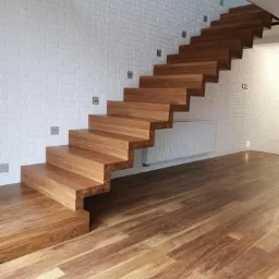 Nowoczesne, drewniane schody wspornikowe z oświetleniem punktowym na białej, ceglanej ścianie, płynnie przechodzące w drewnianą podłogę o podobnym odcieniu.