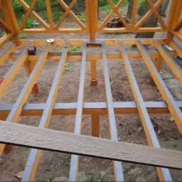 Konstrukcja drewnianej altany w trakcie budowy, z widocznym szkieletem podłogi i fragmentem deski kompozytowej, poziomica na belce.