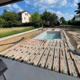 Budowa tarasu wokół basenu: widoczne drewniane legary ułożone na żwirze, przygotowane pod montaż desek tarasowych, w tle dom jednorodzinny i koparka.