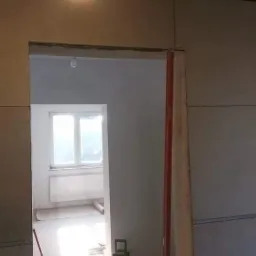 Montaż płyty karton gips na ścianach w trakcie prac wykończeniowych, widoczny fragment sufitu z belkami, surowy stan pomieszczenia z widocznym oknem w tle.