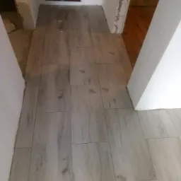 Ułożona jasna podłoga z płytek imitujących drewno w korytarzu, widoczne niedokończone ściany i instalacja grzewcza w tle.