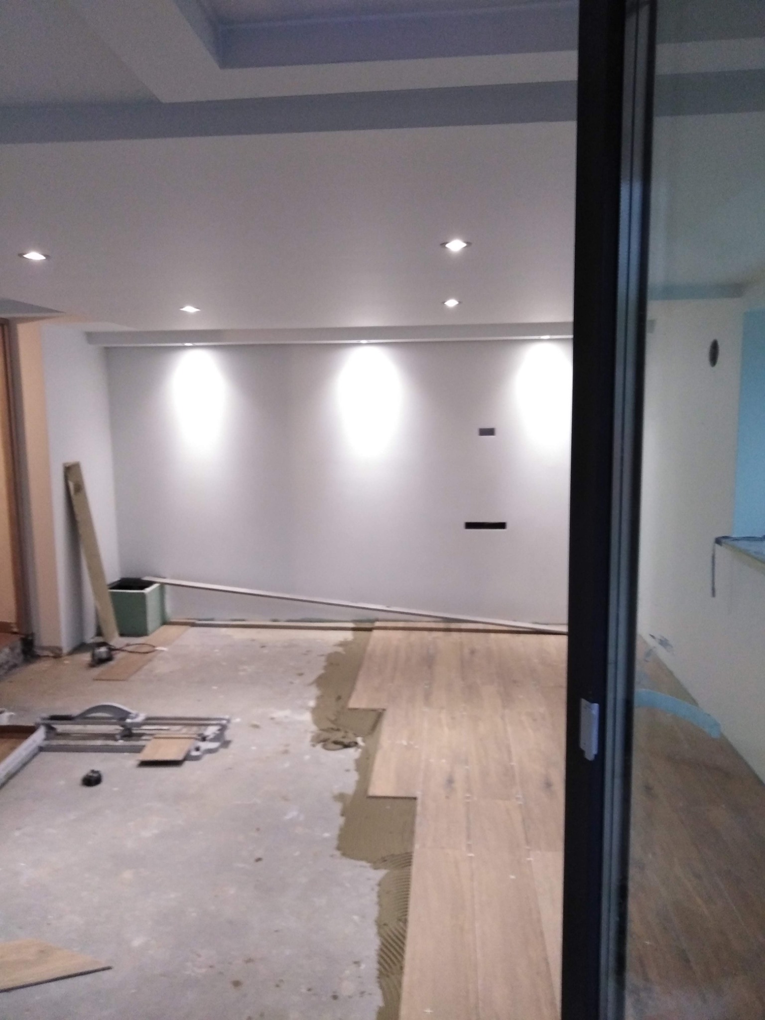 Wnętrze w trakcie układania drewnianej podłogi, widoczny sprzęt do cięcia płytek, surowy beton na podłodze oraz fragment ułożonej podłogi w jasnym odcieniu drewna.