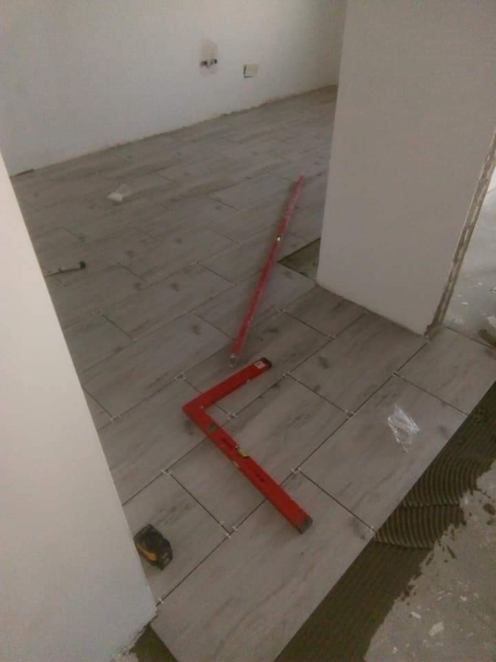 Układanie szarych płytek podłogowych w pomieszczeniu, widoczny poziomica kątowa, poziomica liniowa i miarka przy ścianie.