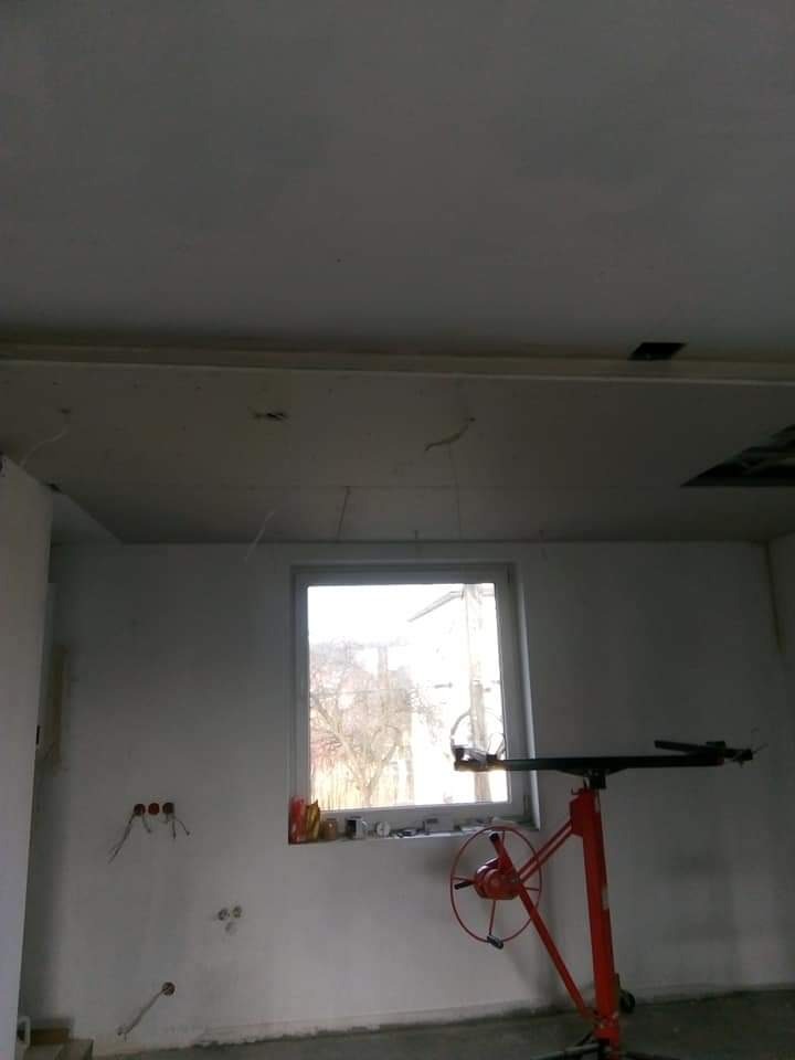Wnętrze w trakcie prac: widoczny podwieszany sufit z płyt kartonowo-gipsowych, czerwony podnośnik do płyt, okno i wystające kable elektryczne ze ściany.