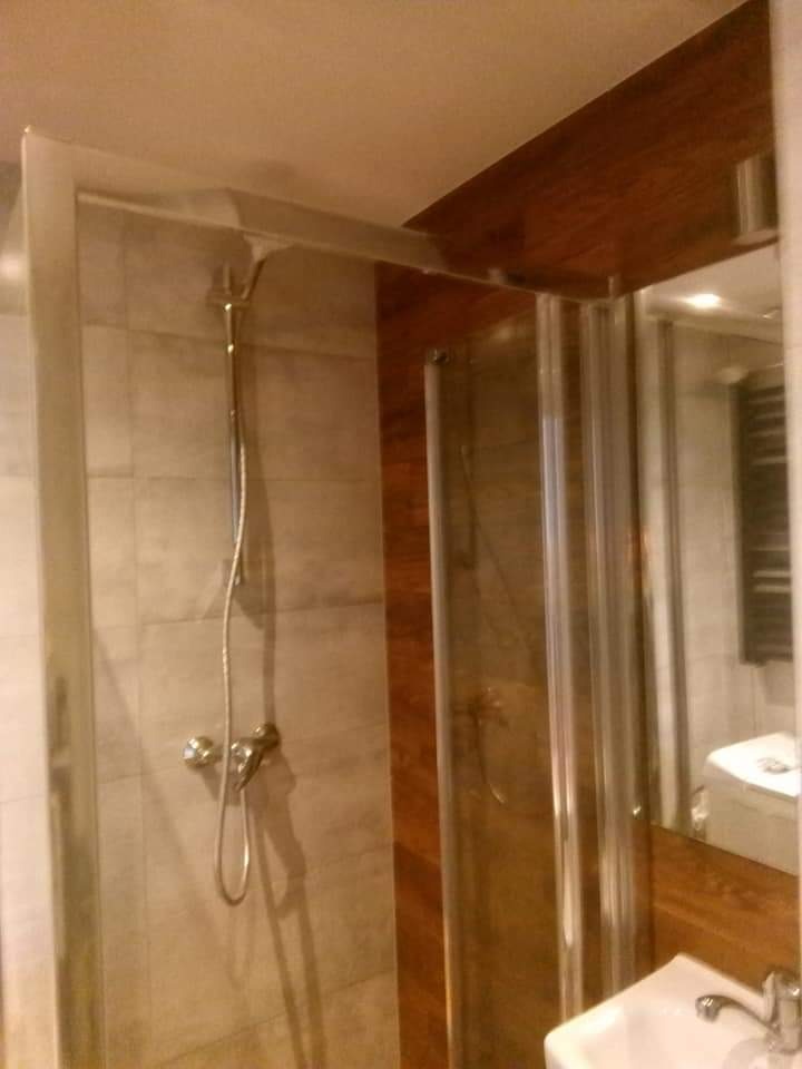 Szklana kabina prysznicowa z chromowaną armaturą na tle szarych płytek i drewnianej boazerii, widok z perspektywy użytkownika.