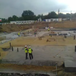 Budowa fundamentów z wystającymi rurami, dwóch pracowników w kamizelkach odblaskowych omawia postęp prac, w tle kontenery budowlane i pochmurne niebo.