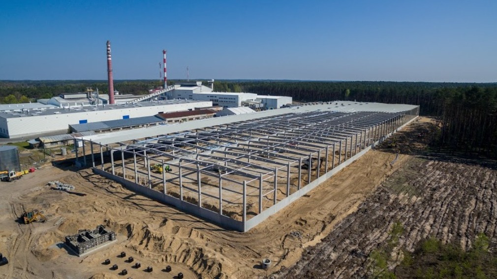 Budowa hali przemysłowej: stalowa konstrukcja szkieletowa z widocznymi słupami i belkami, w tle zabudowania fabryczne z kominami.