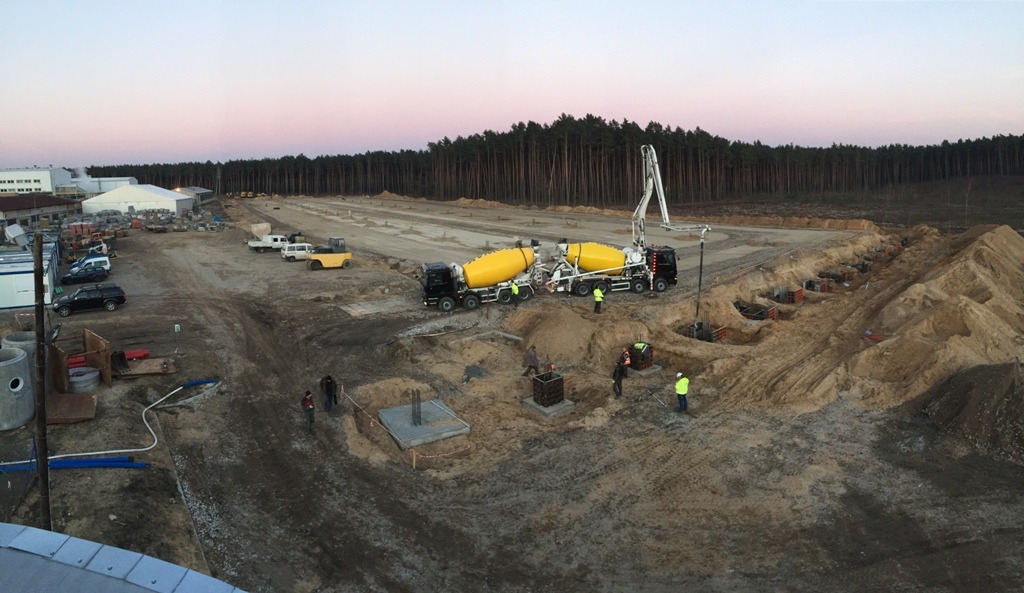 Wylane fundamenty pod budowę hali przemysłowej, widoczne dwa betoniarki z pompami do betonu, pracownicy w kamizelkach odblaskowych, w tle las i budynki.