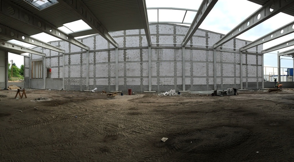 Wnętrze hali w trakcie rozbudowy z widoczną konstrukcją stalową, ścianami z bloczków betonowych i ziemistym podłożem z wyraźnymi śladami opon.
