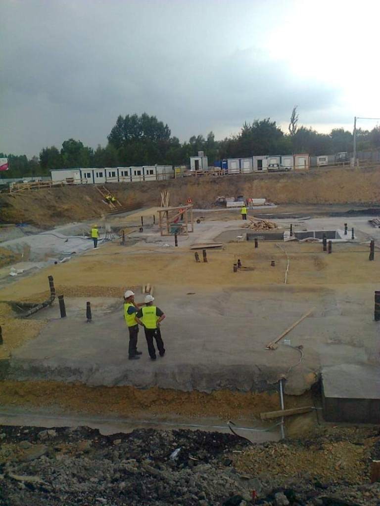 Budowa fundamentów z wystającymi rurami, dwóch pracowników w kamizelkach odblaskowych omawia postęp prac, w tle kontenery budowlane i pochmurne niebo.