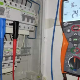 Miernik Sonel MPI-506 wskazujący napięcie 241.1V, podłączony przewodami pomiarowymi do zabezpieczeń w rozdzielnicy elektrycznej Noark.