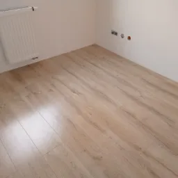 Jasne panele podłogowe ułożone w rogu pokoju z białymi ścianami, widoczny grzejnik i niezamaskowane gniazdka elektryczne.