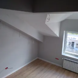 Pokój na poddaszu po malowaniu ścian na szaro, z drewnianą podłogą, grzejnikiem pod oknem i wystającymi kablami elektrycznymi ze ściany i sufitu.