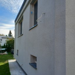 RemontTech-M Mariusz Zych - Świeżo pomalowana elewacja domu w odcieniach szarości, widoczna różnica w kolorze na ścianie, z oknami i rurami na zewnątrz, trawnik w tle.