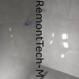 RemontTech-M Mariusz Zych - Świeżo ułożone, szare płytki podłogowe w łazience, z widocznym odpływem liniowym i elementami instalacji wodnej wystającymi ze ściany, zabezpieczonymi niebieską folią.