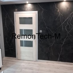 RemontTech-M Mariusz Zych - Czarna ściana z efektem marmuru i białymi żyłkami, jasne drzwi z szybami, podłoga z paneli imitujących drewno.