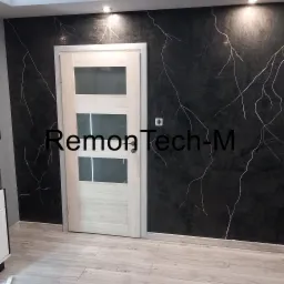 Czarna ściana z efektem marmuru i białymi żyłkami, jasne drzwi z szybami, podłoga z paneli imitujących drewno.