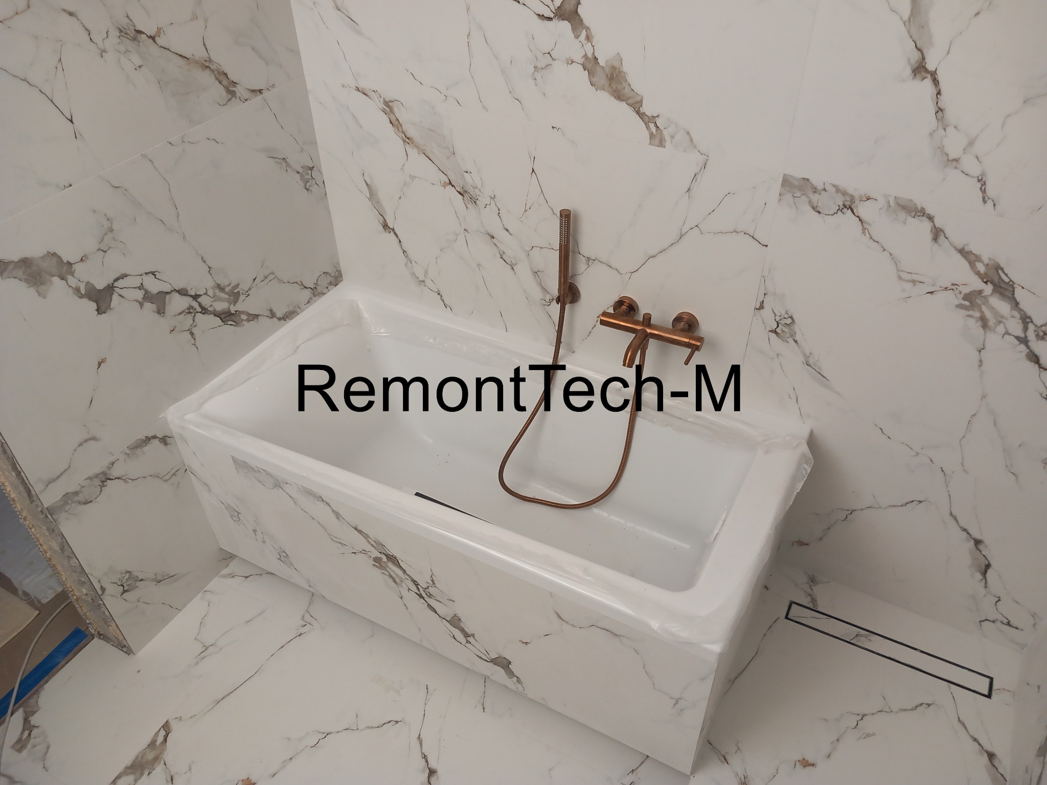 Biała wanna z marmurowym wzorem, złota armatura, odpływ liniowy. Całość w jasnej łazience, wykończonej płytkami imitującymi marmur.