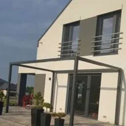 Nowoczesna pergola o prostej, geometrycznej konstrukcji z ciemnego metalu, przylegająca do białej ściany domu z dużymi oknami i metalowymi balustradami. Widoczny fragment tarasu wyłożonego szarą...
