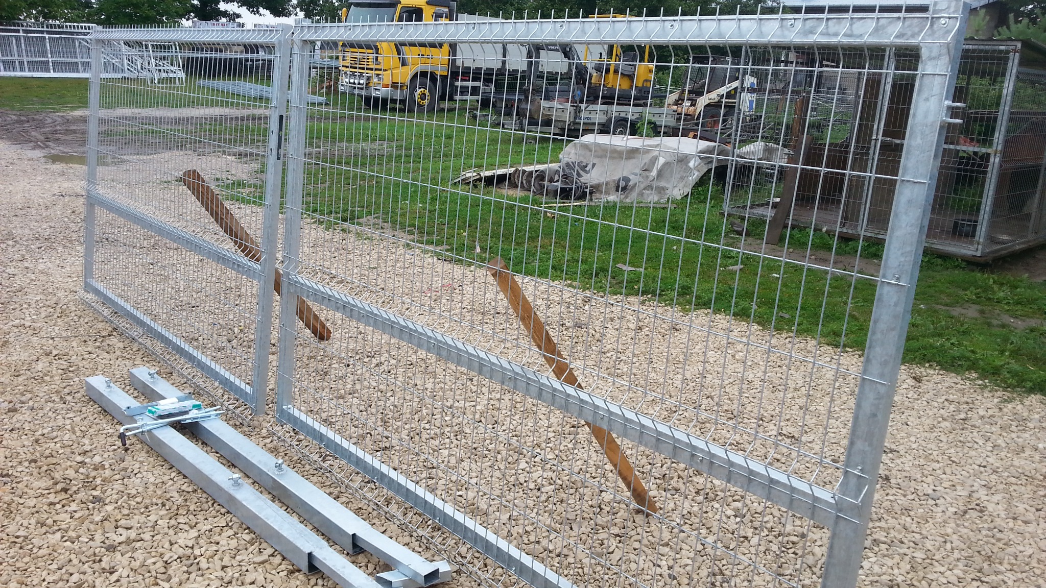 Fragment montażu ocynkowanej bramy z siatki panelowej, widoczne elementy mocujące, podpory drewniane i pojazdy budowlane w tle.