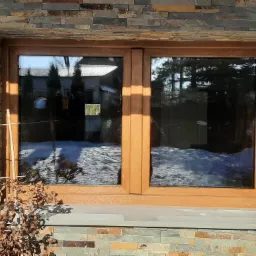 Dwuskrzydłowe okno z drewnianą ramą, zamontowane w elewacji z kamienia łupanego; widoczny śnieg za oknem i odbicie zabudowań.