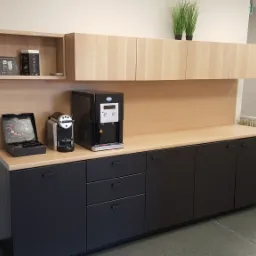 Kącik biurowy z dystrybutorem wody, ekspresem do kawy i szafkami w odcieniach beżu i czerni, uzupełniony o rośliny doniczkowe na górnych szafkach.