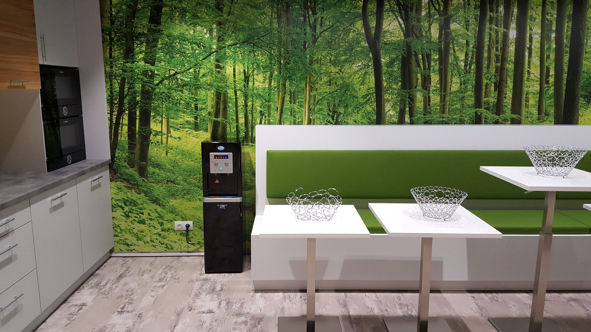 Nowoczesny dystrybutor wody w biurze, z zielonym fototapetą lasu w tle, obok białe szafki kuchenne i stoliki z dekoracyjnymi koszyczkami.