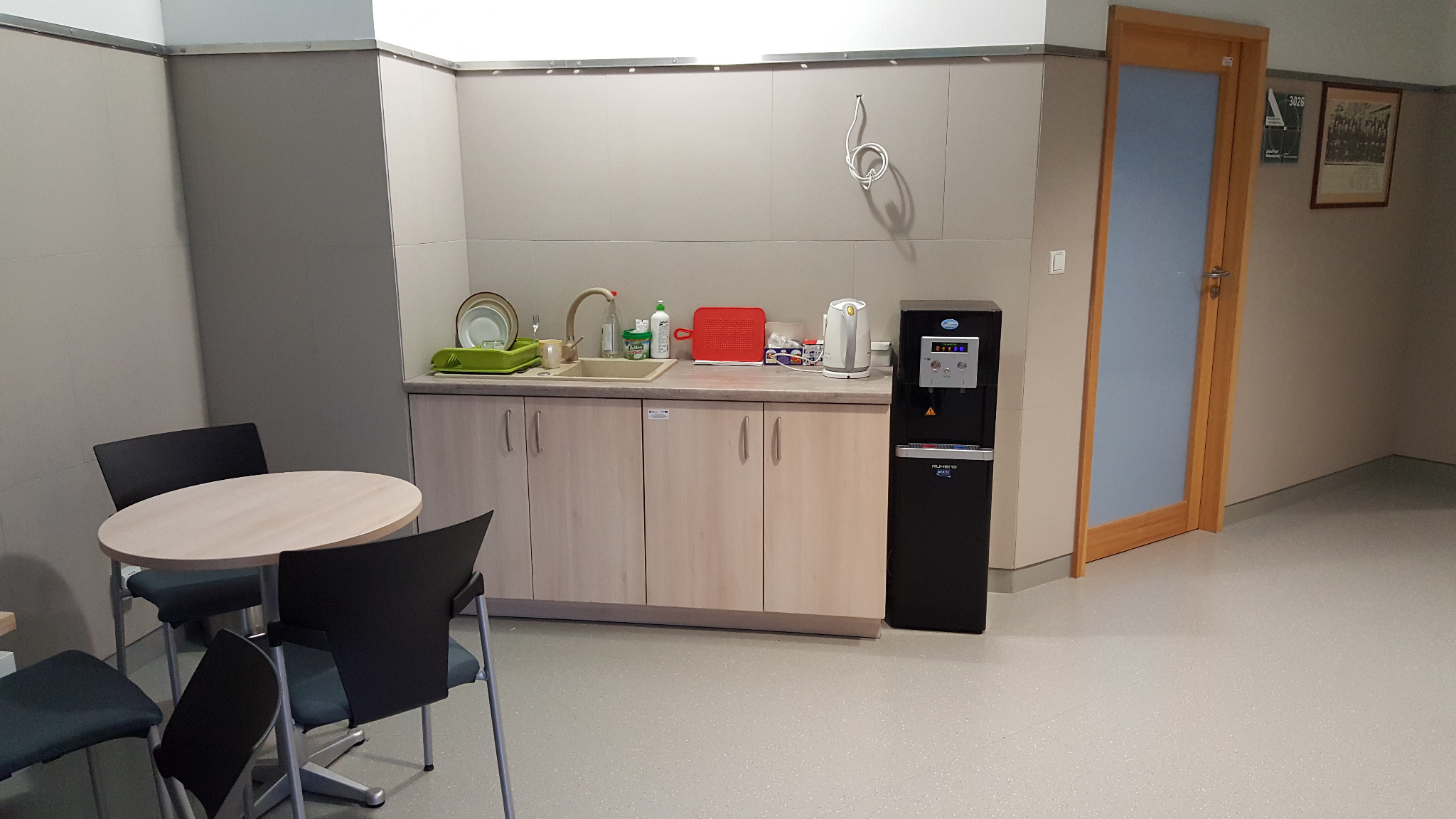 Kącik socjalny w biurze firmy z Nur: czarny dystrybutor wody obok szafek kuchennych, na blacie naczynia, czajnik i artykuły spożywcze, obok okrągły stół z krzesłami.