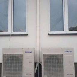 Dwa identyczne, białe klimatyzatory marki Samsung zamontowane na jasnej elewacji budynku pod dwoma oknami. Widoczne rury odprowadzające skropliny. Minimalistyczny design.