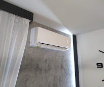 Biały klimatyzator ścienny zamontowany w nowoczesnym wnętrzu z szarą ścianą o fakturze betonu i białymi zasłonami.