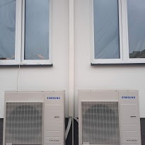 Dwa identyczne, białe klimatyzatory marki Samsung zamontowane na jasnej elewacji budynku pod dwoma oknami. Widoczne rury odprowadzające skropliny. Minimalistyczny design.