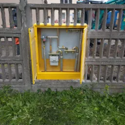 Żółta szafka gazowa z otwartymi drzwiczkami, zamontowana w betonowym ogrodzeniu, widoczny gazomierz i rury, w tle wykop i maszyna budowlana.