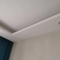 Wykończony sufit z płyt kartonowo-gipsowych z wbudowanym oświetleniem punktowym i fragmentem zasłony w kolorze butelkowej zieleni.