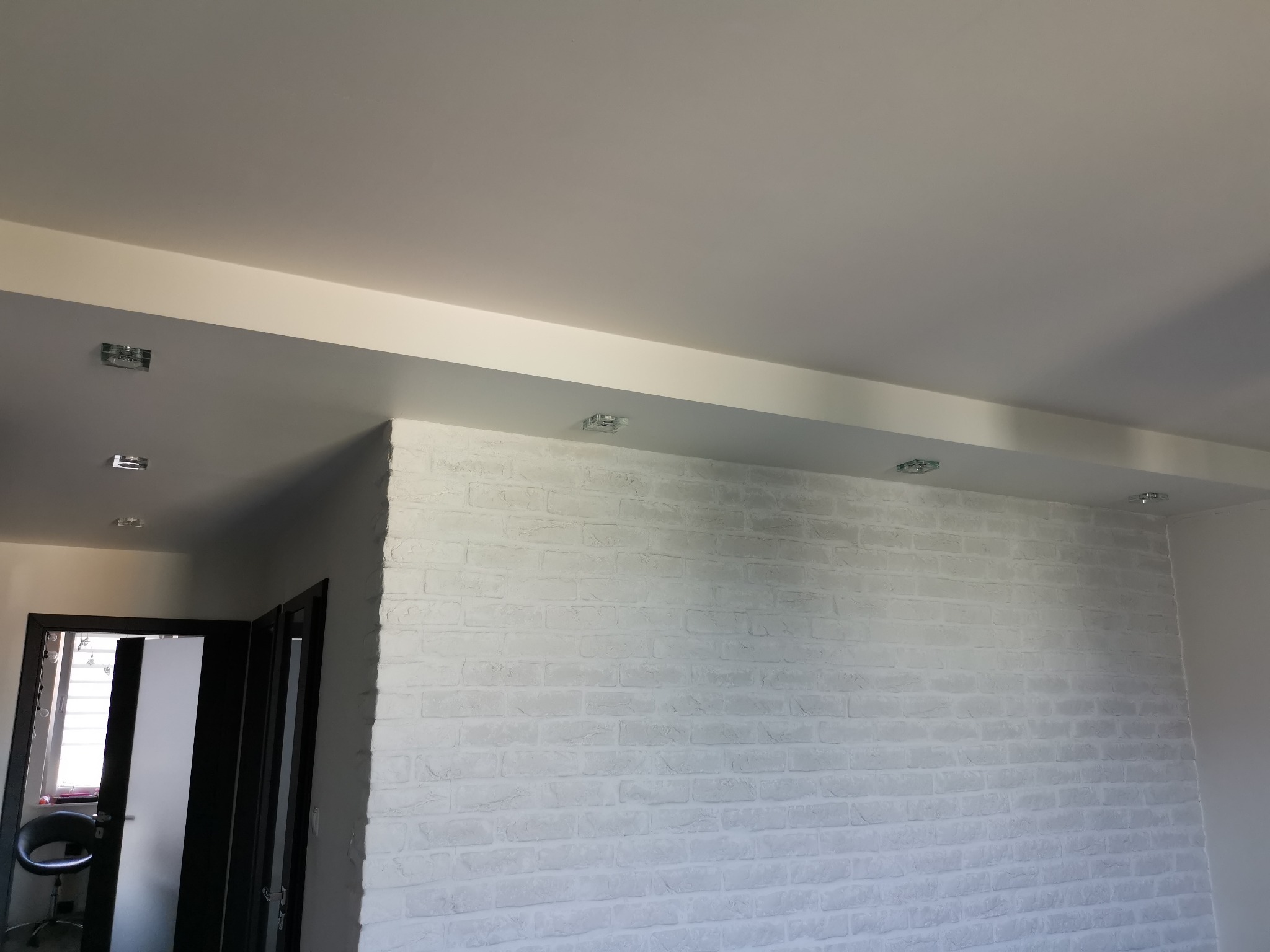 Wnętrze z białą ścianą imitującą cegłę, oświetlenie punktowe w suficie podwieszanym, widoczne fragmenty drzwi w ciemnej ościeżnicy.
