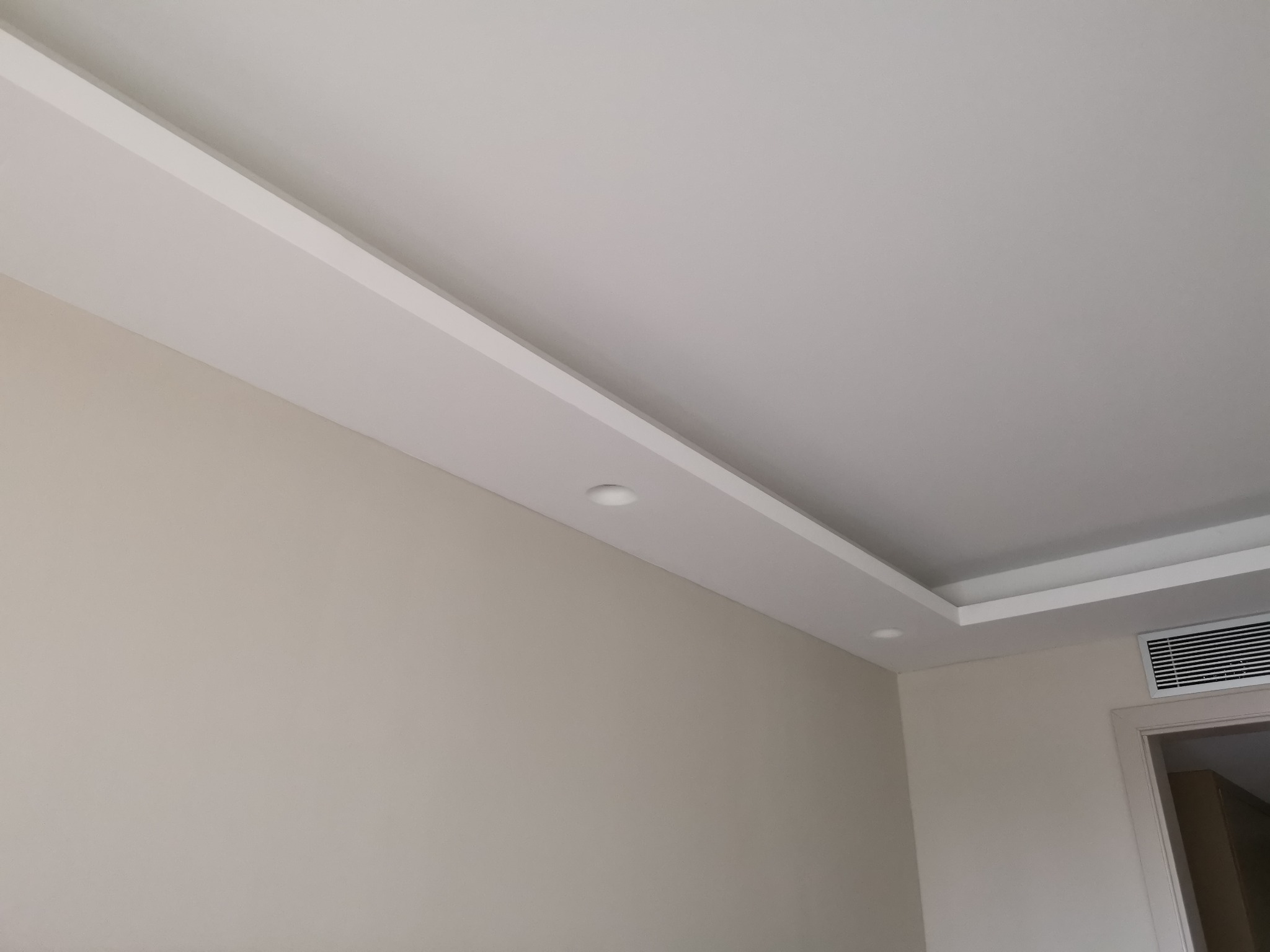 Sufit podwieszany z oświetleniem punktowym, widoczna ściana w odcieniach beżu i fragment klimatyzacji przy drzwiach.