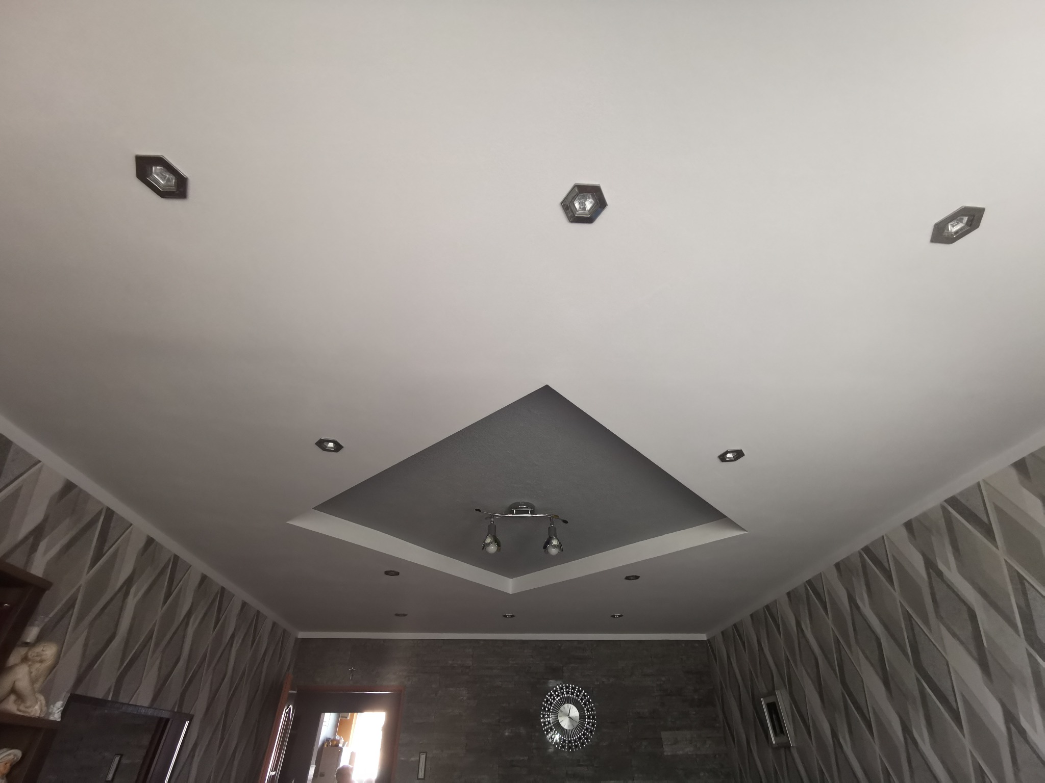 Wykończony sufit z dekoracyjną wnęką w kształcie rombu, oświetleniem punktowym i centralną lampą z dwoma reflektorami, ściany pokryte tapetą w geometryczny wzór.