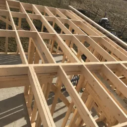 Konstrukcja drewniana dachu w trakcie budowy, widok z góry na szkielet z belek i desek, tło stanowią pola i łąki.