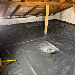 Strop betonowy poddasza zabezpieczony czarną folią, widoczne ślady butów, drewniana konstrukcja dachu oraz lampa budowlana oświetlająca przestrzeń.