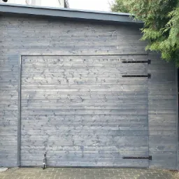 Szary drewniany garaż z poziomych desek z czarnymi zawiasami, przylegający do białego budynku, z zielonymi krzewami po prawej stronie.