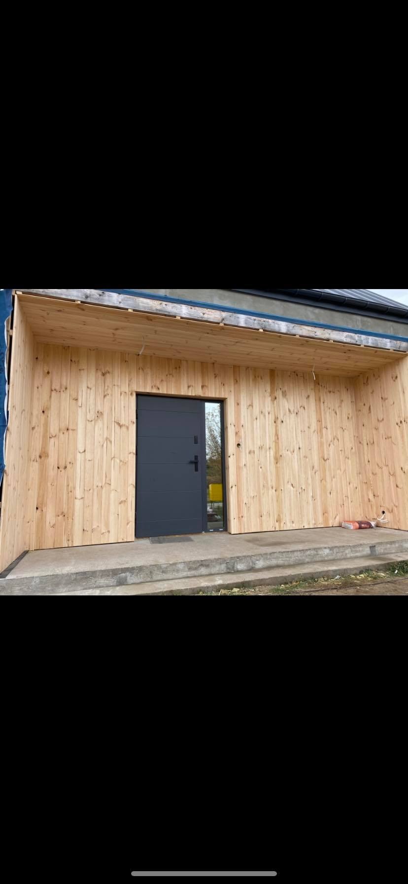 Drewniana elewacja budynku z szarą, nowoczesną stolarką drzwiową i zadaszeniem, widoczny fragment trawnika i schody wejściowe.