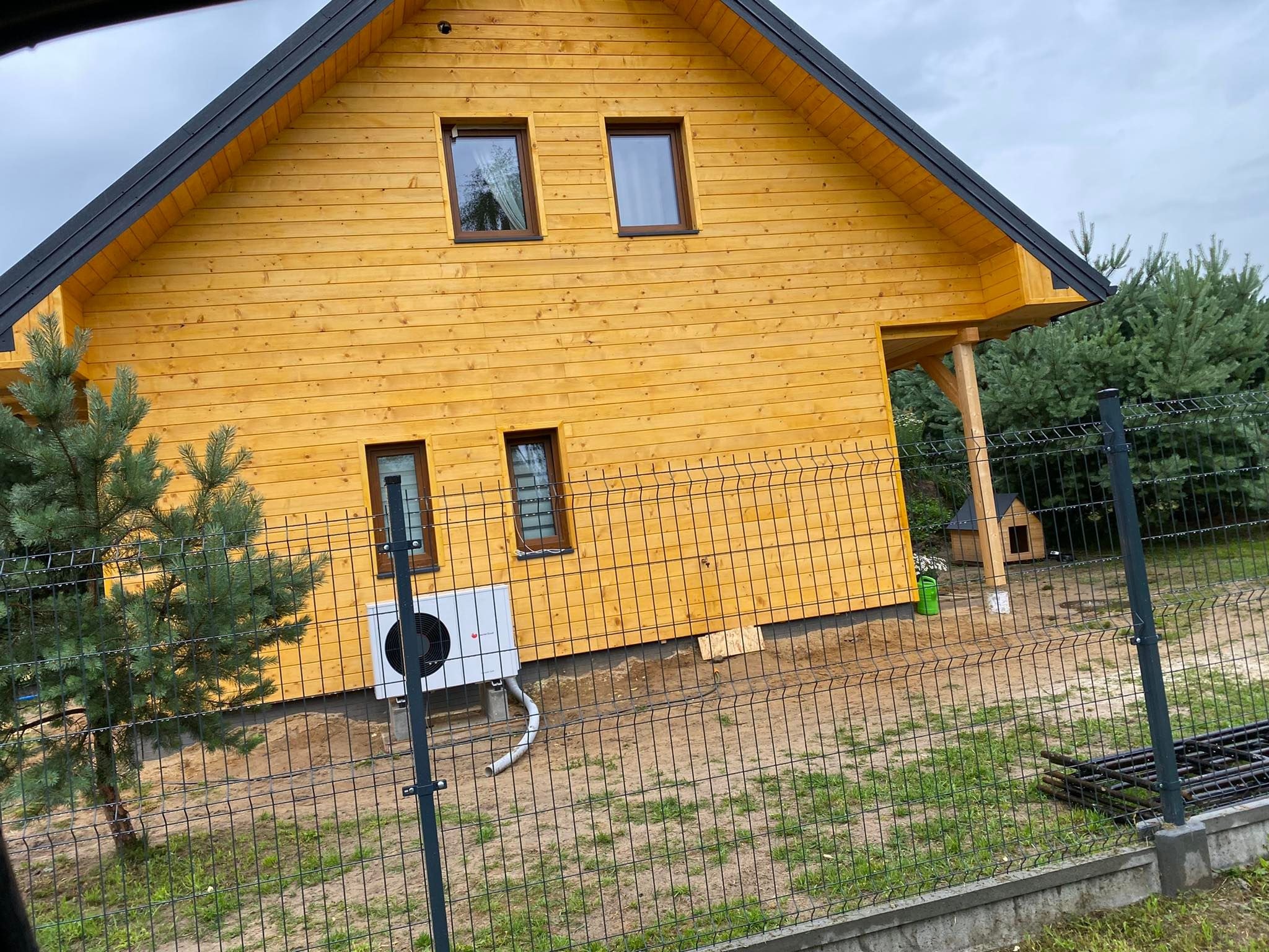 Nowo wybudowany, żółty dom z drewnianą elewacją, widoczny zza ogrodzenia z siatki, z zamontowaną klimatyzacją na bocznej ścianie i budą dla psa w tle.