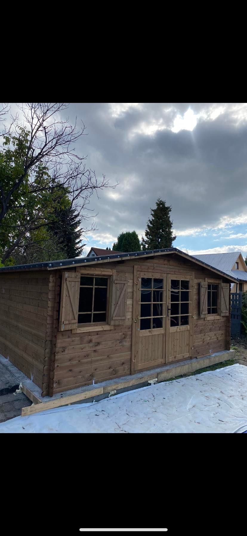 Nowa drewniana konstrukcja z oknami i drzwiami, prawdopodobnie domek narzędziowy, na fundamencie zabezpieczonym folią, w tle drzewa i domy pod pochmurnym niebem.