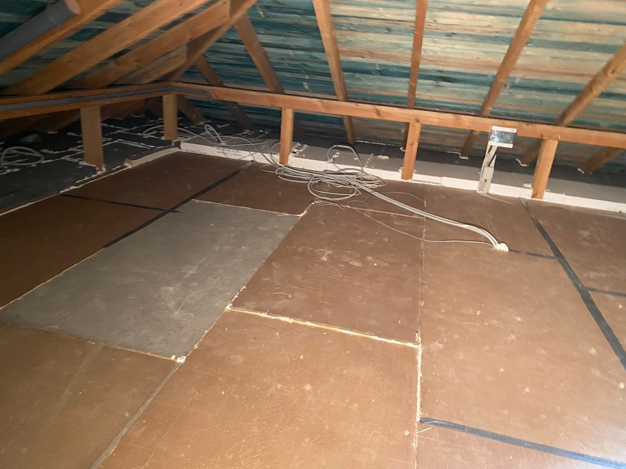 Strop betonowy poddasza z widoczną izolacją piankową przy ścianach, częściowo pokryty brązowymi płytami podłogowymi, z luźno leżącymi kablami elektrycznymi i widoczną konstrukcją dachu.