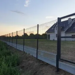 Długa linia nowoczesnego ogrodzenia z paneli drucianych na betonowej podmurówce, z otwartą furtką, na tle domu i zachodzącego słońca.