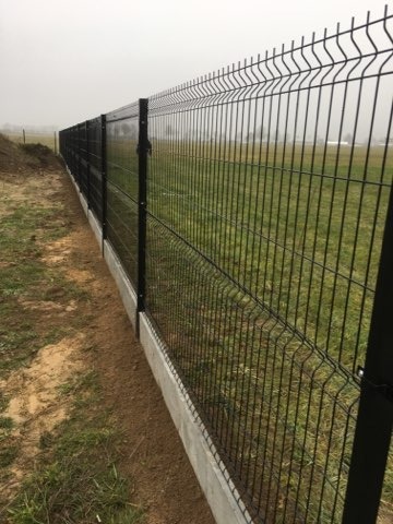 Długa, czarna, metalowa konstrukcja z siatki na betonowej podmurówce, oddzielająca teren zielony od drogi gruntowej w pochmurny dzień.