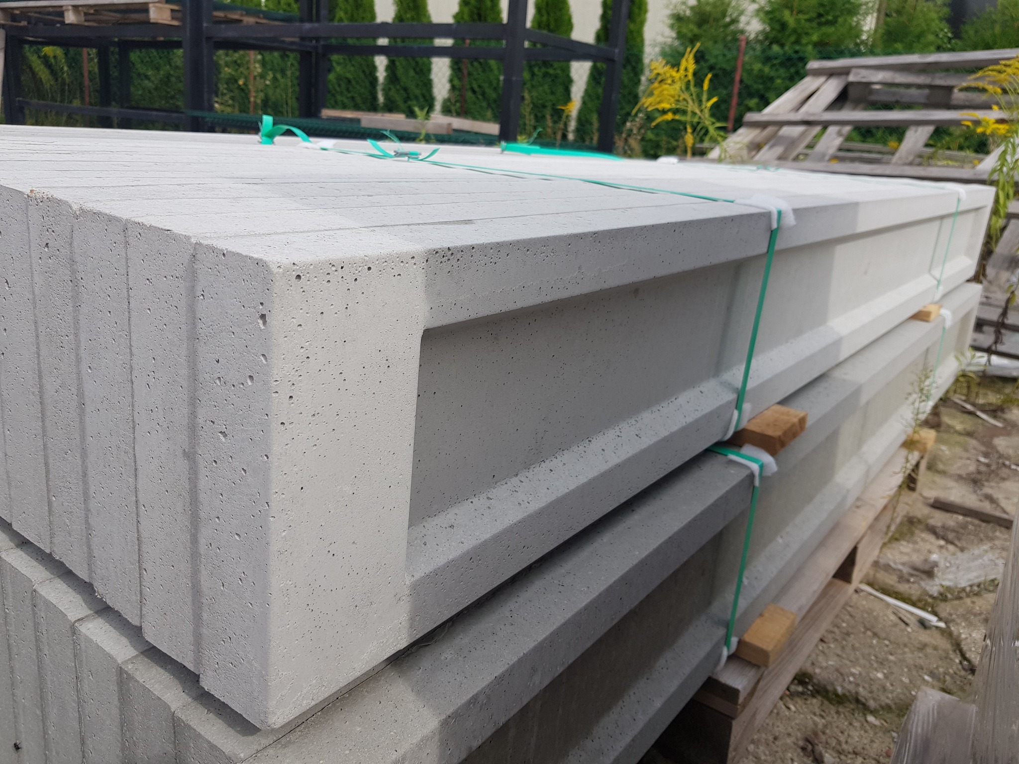 Szare betonowe prefabrykaty ogrodzeniowe typu L, ułożone na palecie, zabezpieczone zieloną taśmą, widok z bliska na fakturę betonu i kształt elementów.