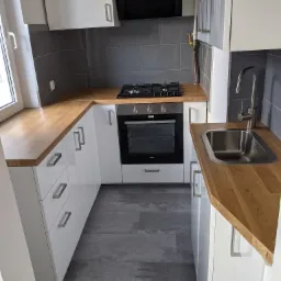 Nowoczesna kuchnia w odcieniach bieli i szarości z drewnianym blatem, szare płytki na ścianie, widoczna kuchenka gazowa i piekarnik Amica, szare panele podłogowe.