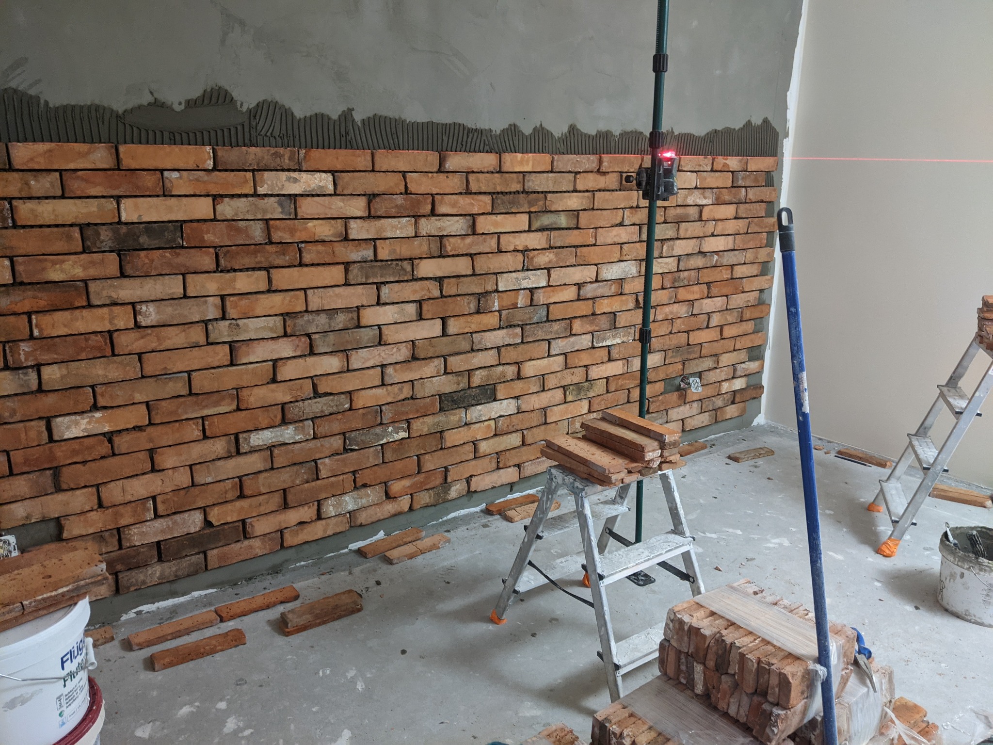 Montaż dekoracyjnej ściany z cegieł: widoczny fragment ściany z nałożonym klejem, częściowo ułożone cegły, poziomica laserowa, drabina i materiały budowlane na podłodze.