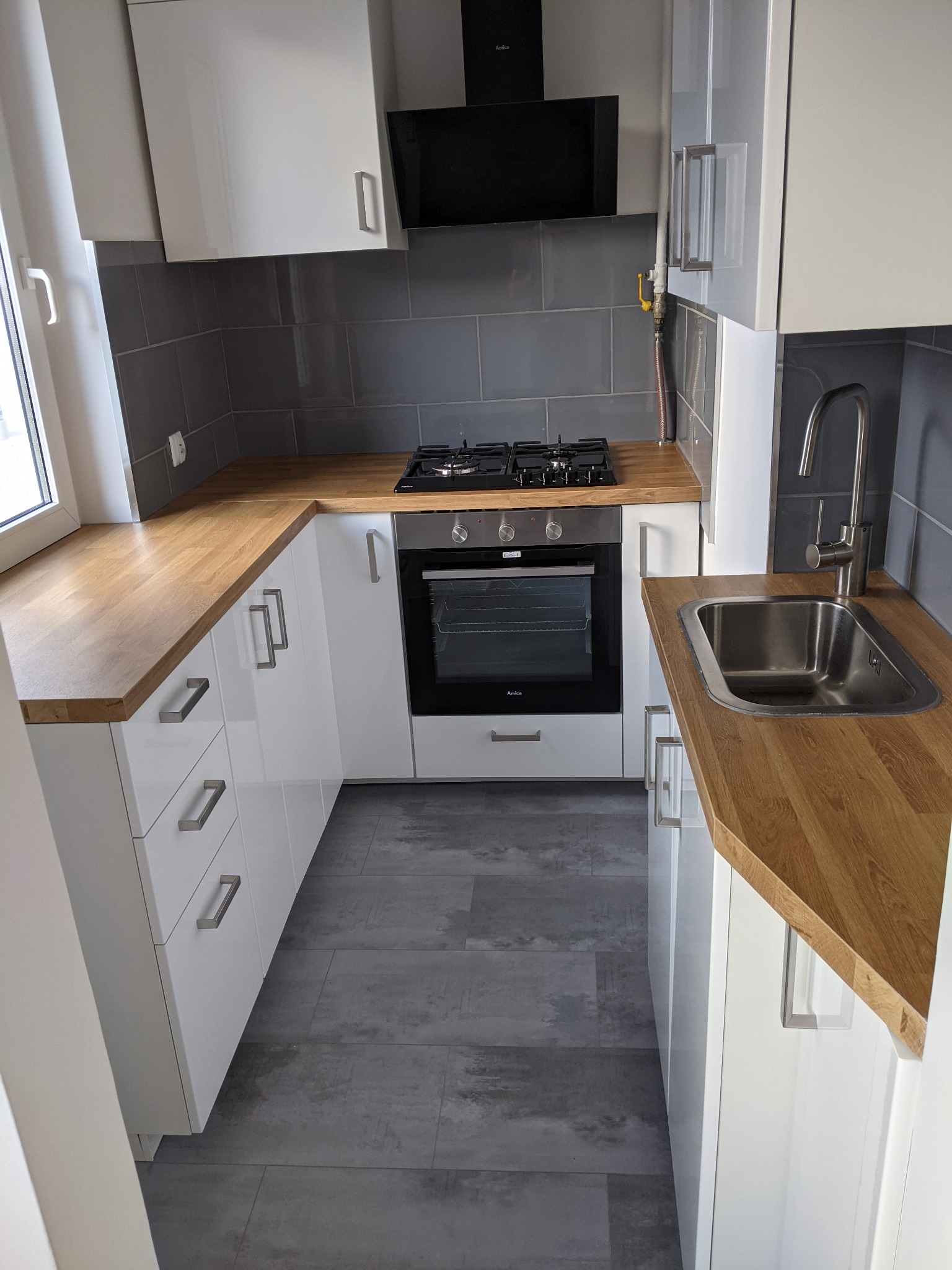 Nowoczesna kuchnia w odcieniach bieli i szarości z drewnianym blatem, szare płytki na ścianie, widoczna kuchenka gazowa i piekarnik Amica, szare panele podłogowe.